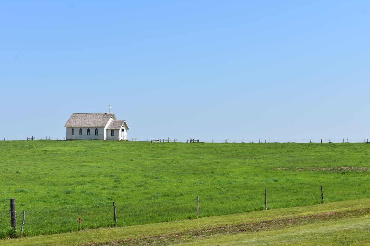 A solitary rural church stands peacefully amidst an expansive green field under a clear blue sky.